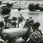 American troops landing at Omaha Beach, June 6, 1944. The presence of tracked vehicles and DUKWs (“Ducks”) on the beach and the organized columns of men heading inland indicate that this photo was taken several hours after the initial infantry assault.